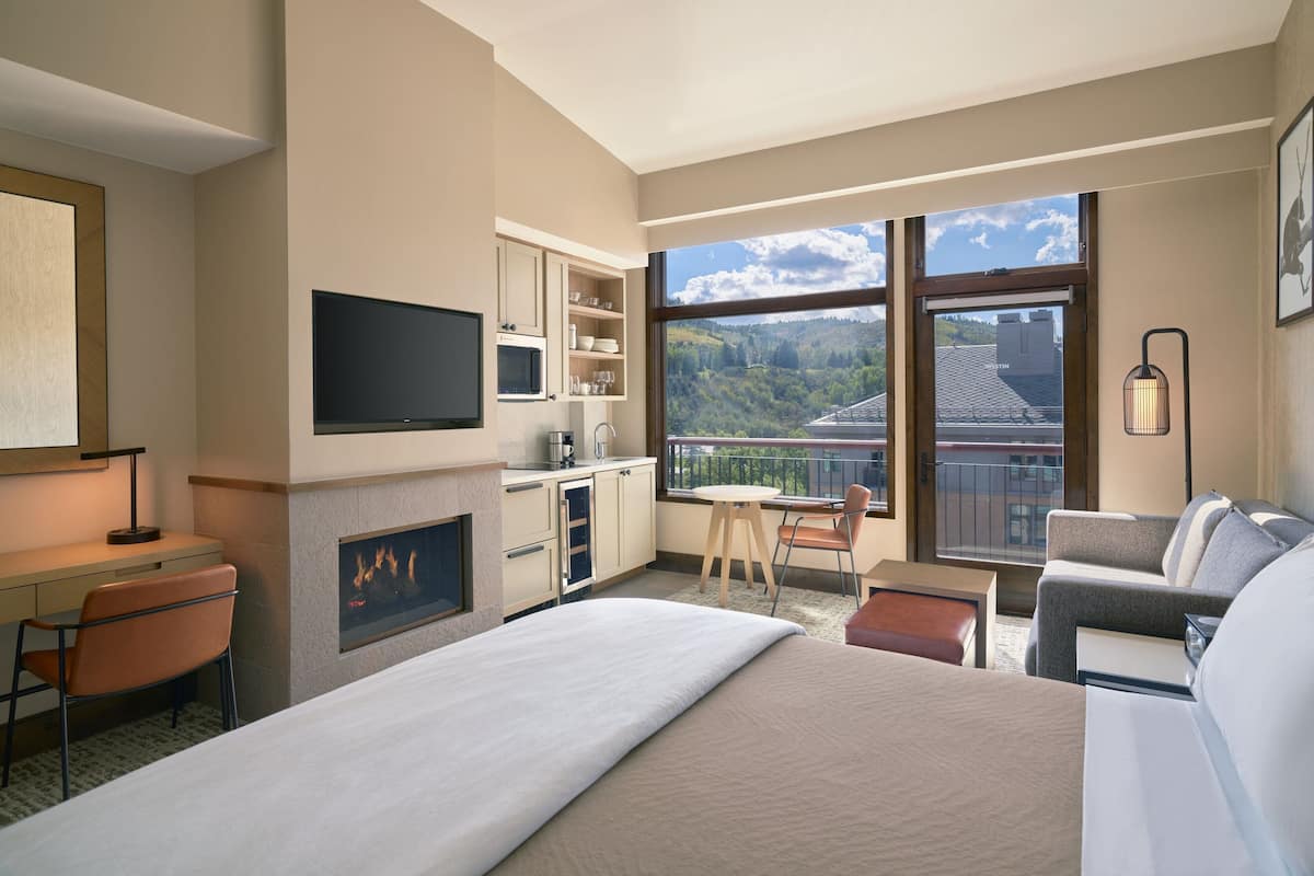 A modern hotel room with a large bed, a desk, a TV, a fireplace, a balcony with a view of the mountains, and a sofa.