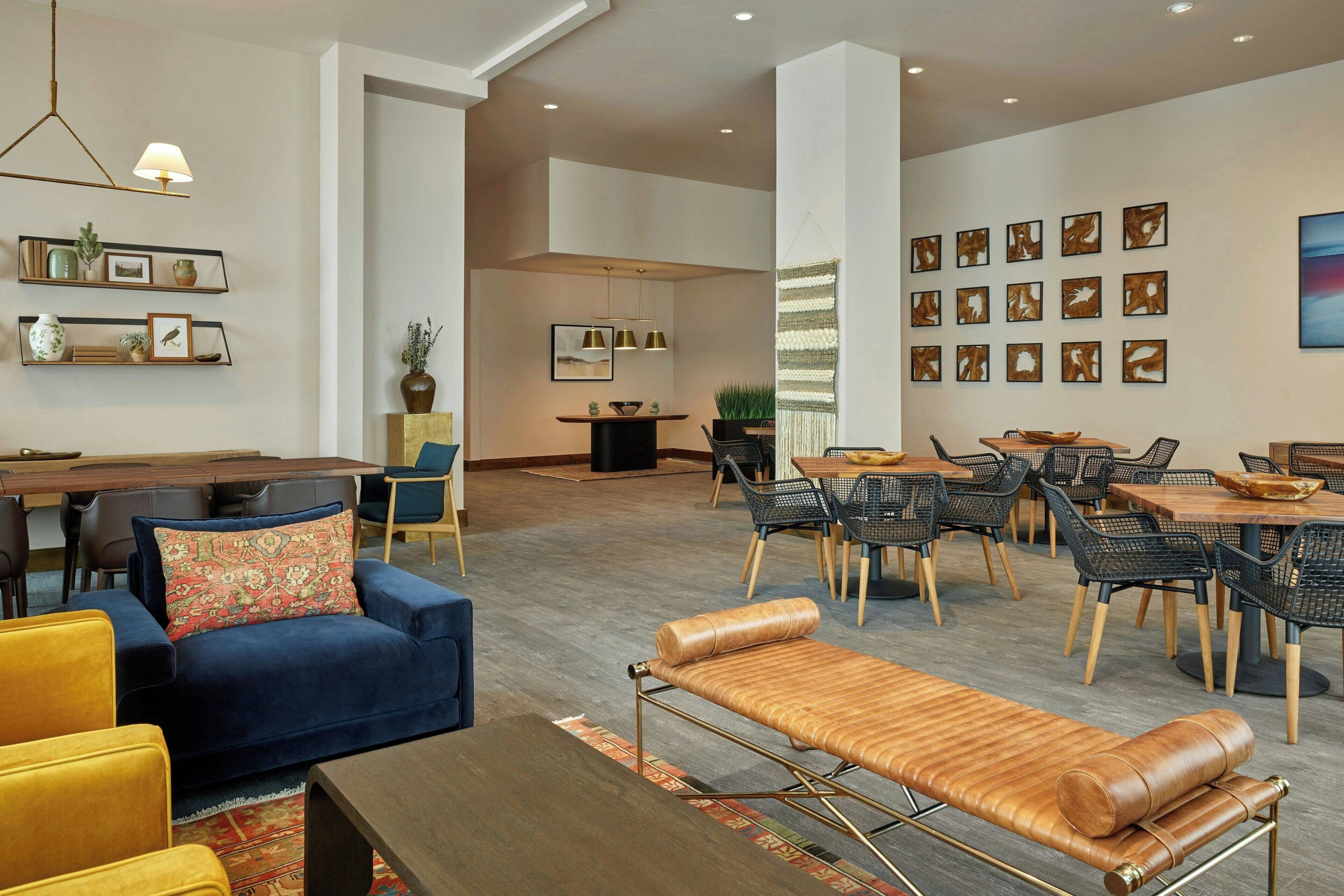 A modern hotel lobby with a blue sofa, yellow chairs, a wooden bench, and a dining area with tables and chairs.