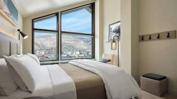 A hotel room with a large window, a bed, a chair, a bedside table, and a wall-mounted lamp.