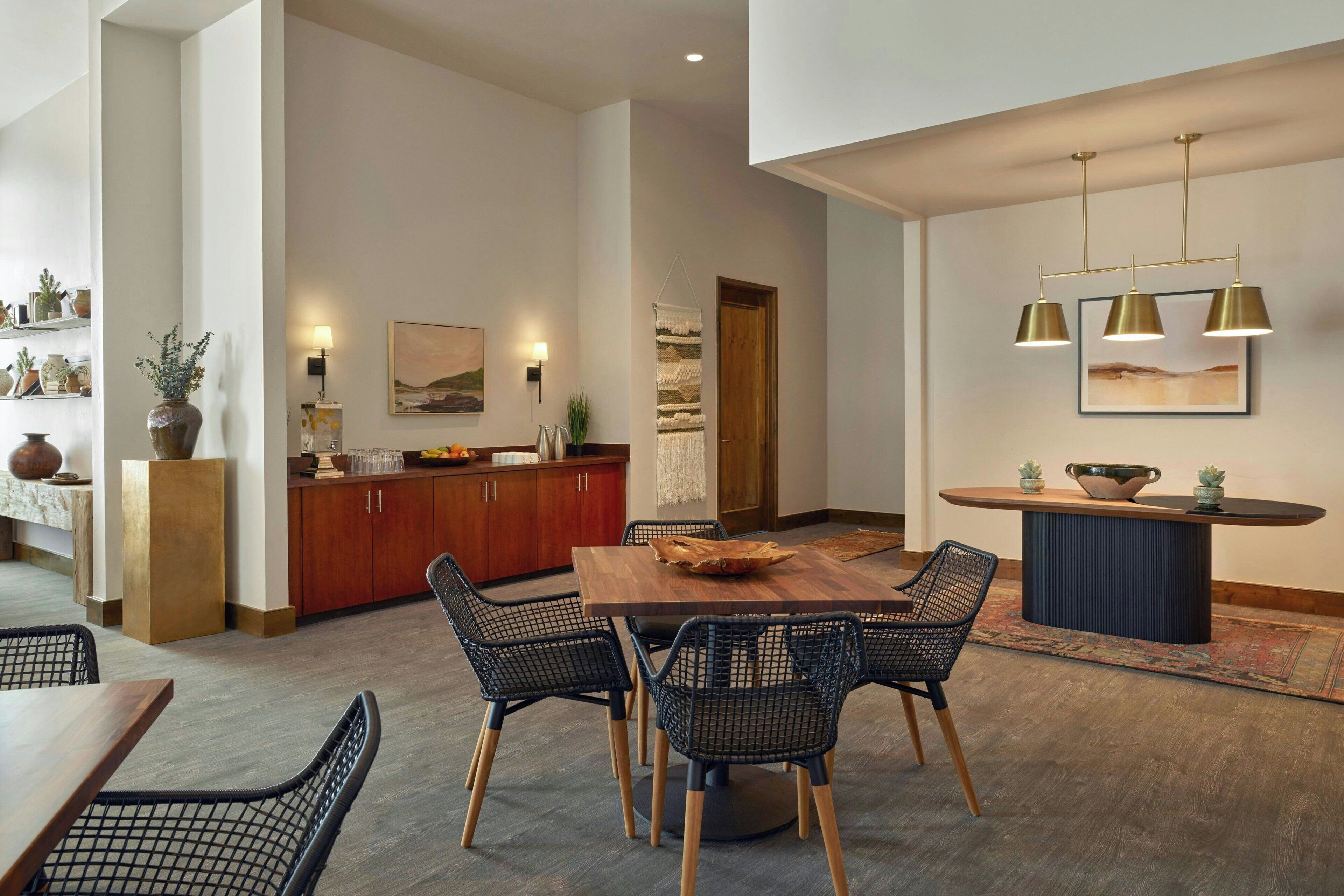A modern dining area with a wooden table and black wireframe chairs, a kitchen with wooden cabinets, and pendant lighting.