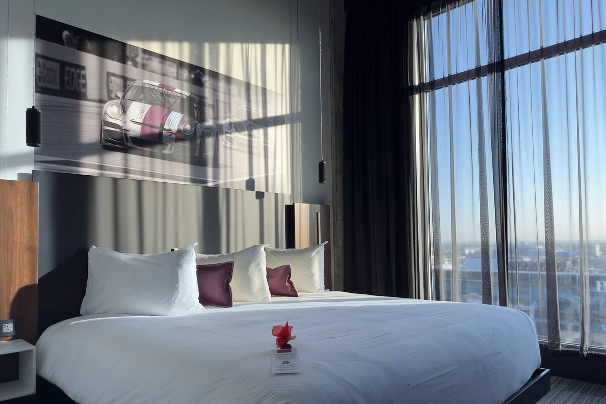 A modern hotel room with a large bed, a nightstand, a wall-mounted TV displaying a racing car, and a city view through large windows.
