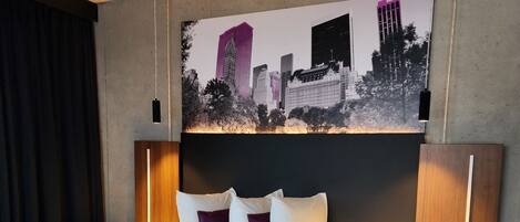 Une chambre d’hôtel moderne avec un grand lit, une œuvre d’art représentant un paysage urbain accrochée au mur et une petite table de chevet avec une lampe.