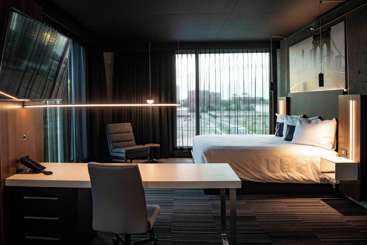 Habitación de hotel con cama, escritorio, silla y un ventanal con vistas.