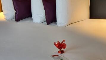 Un lit bien fait, avec des oreillers blancs et violets, un objet décoratif rouge et une petite carte posée sur le lit.