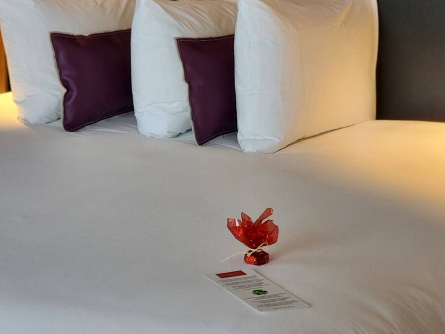 Un lit bien fait, avec des oreillers blancs et violets, un objet décoratif rouge et une petite carte posée sur le lit.
