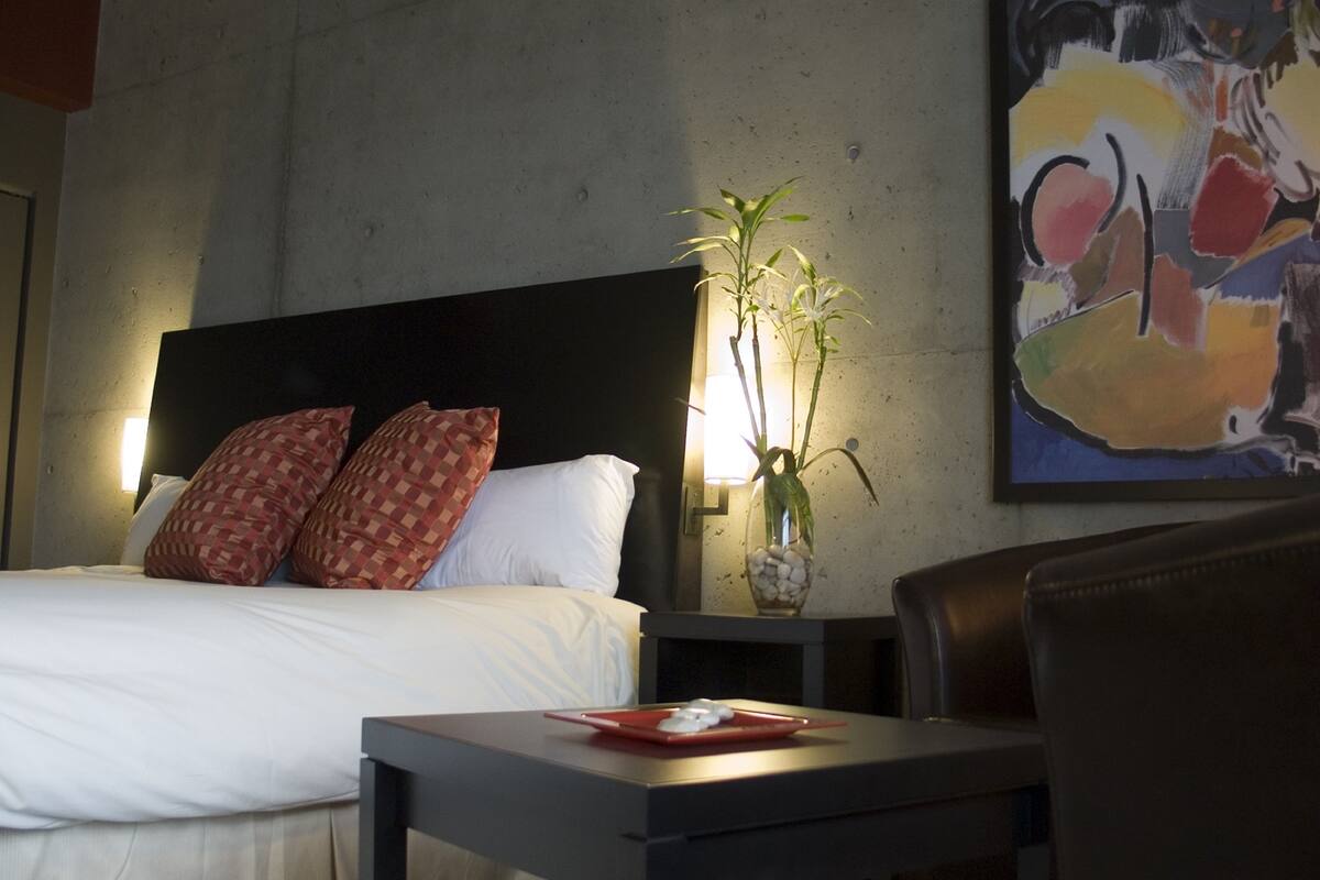 A hotel room with a bed, a nightstand, a table, a chair, a lamp, and a painting on the wall.