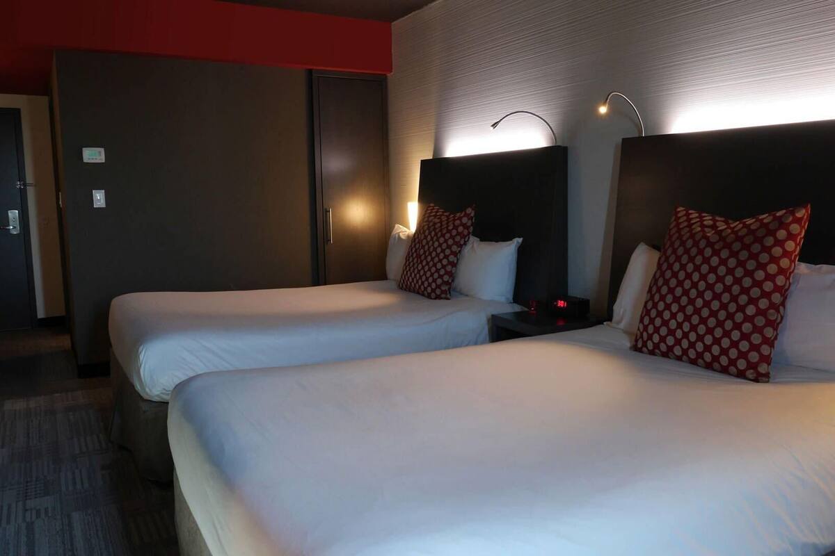 A hotel room with two beds, a nightstand, a wall-mounted lamp, and a door.