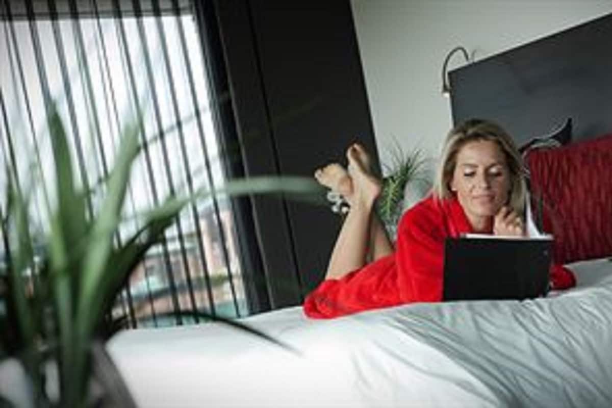 A person lying on a bed with a book, in a room with a window and a plant.