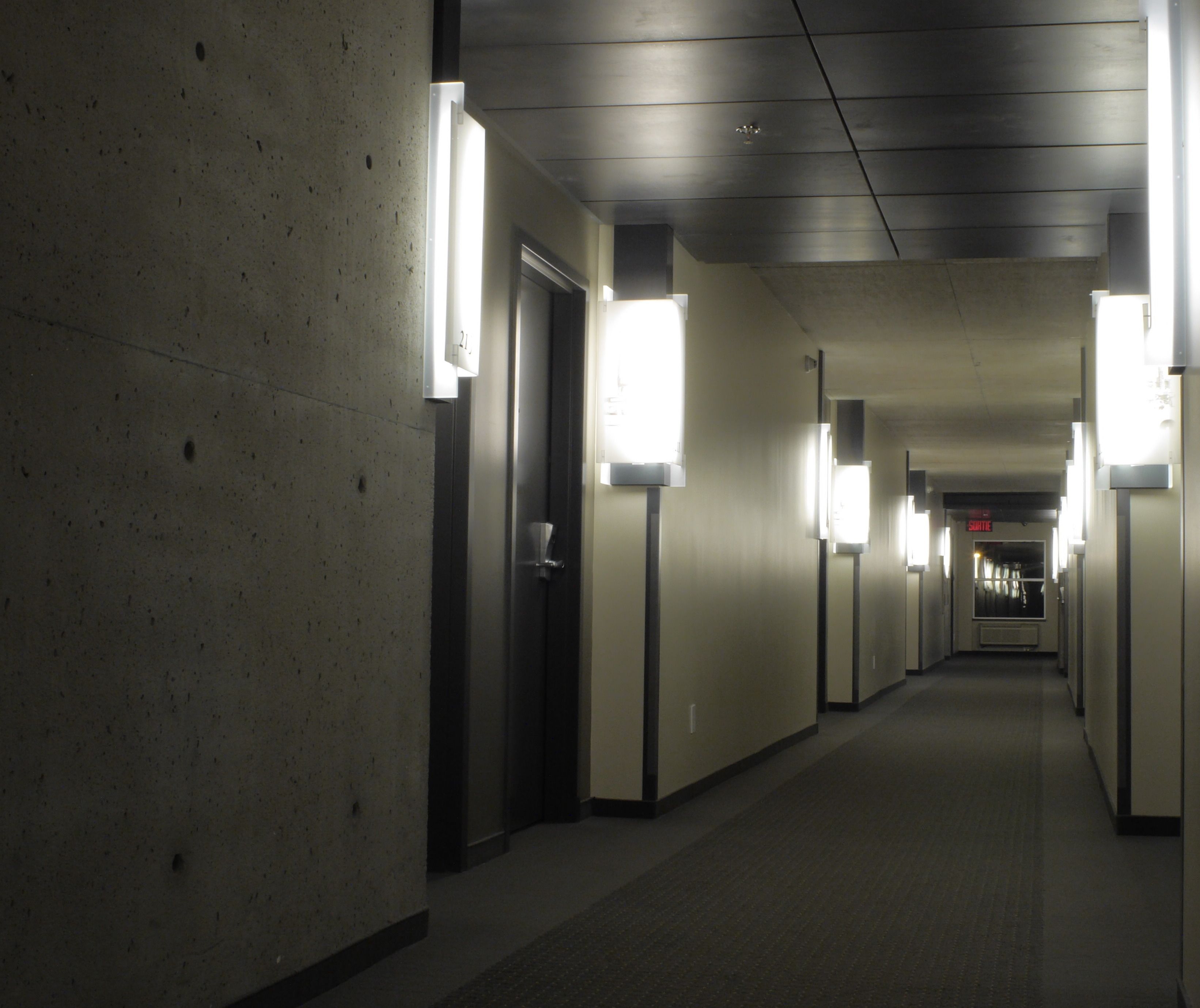 Un pasillo largo y estrecho con iluminación fluorescente, paredes de concreto y puertas a ambos lados.