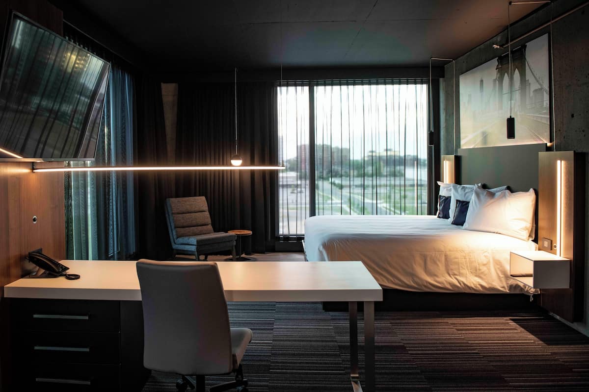 A hotel room with a bed, desk, chair, and a large window with a view of a bridge.