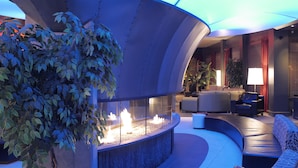 Un área de lounge moderna con una disposición curva de asientos, una chimenea central y una gran estructura parecida a una sombrilla que proporciona sombra.