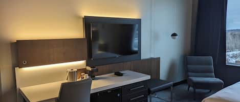 A modern hotel room with a flat-screen TV mounted on the wall, a desk with a chair, a bench, and a bed.