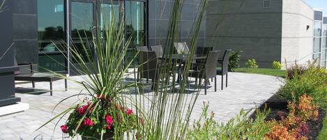 Un área de estar al aire libre con sillas y mesas, rodeada de plantas en macetas y un edificio moderno con amplias ventanas al fondo.