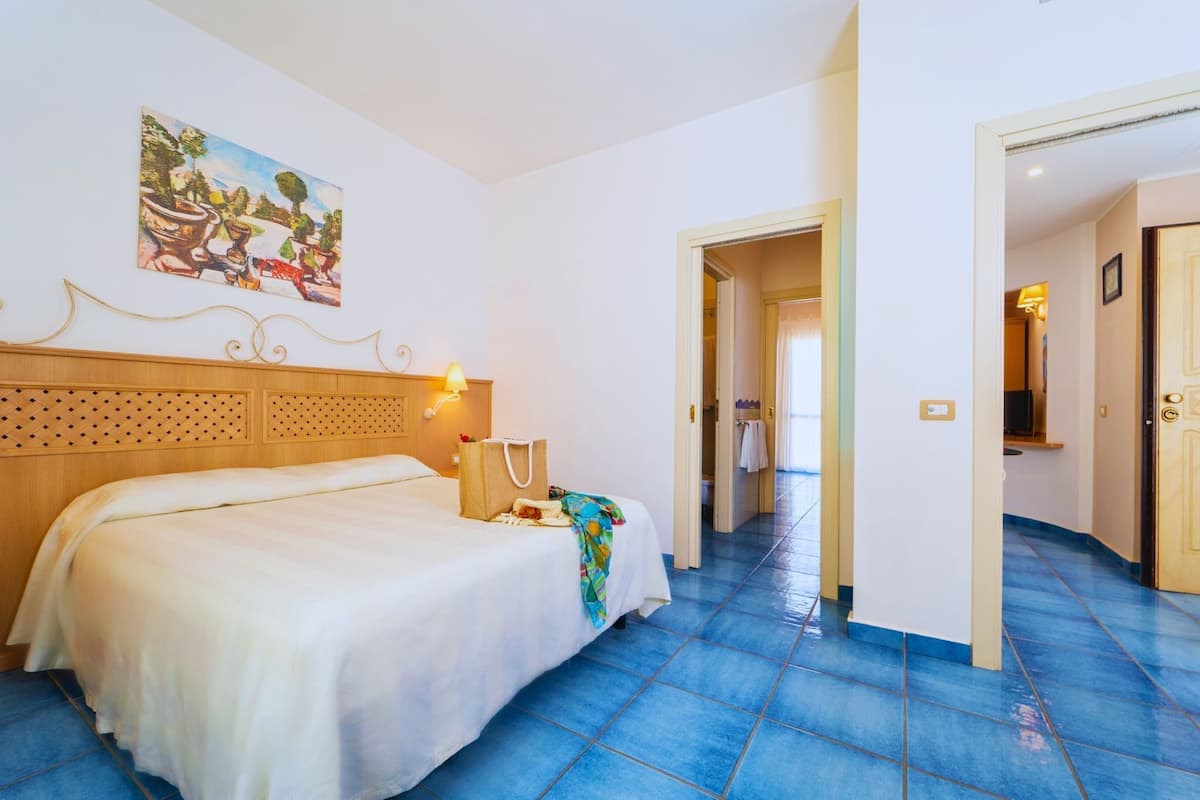 A hotel room with a bed, a painting on the wall, a wooden headboard, a nightstand with a lamp, and a blue tiled floor.