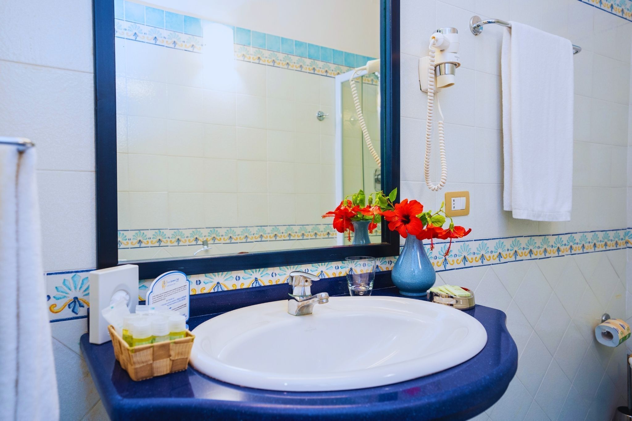 A bathroom with a white sink, blue countertop, and a mirror. There is a vase with red flowers on the counter.