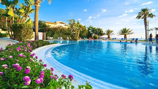 Una piscina rodeada de palmeras y sillas de descanso, con un cielo azul despejado al fondo.