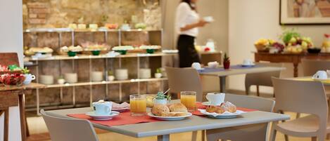 A breakfast buffet with various food items and beverages, a television screen in the background, and a woman serving.