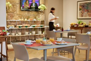 Un bufé de desayuno con diferentes alimentos y bebidas, una pantalla de televisión al fondo y una camarera.