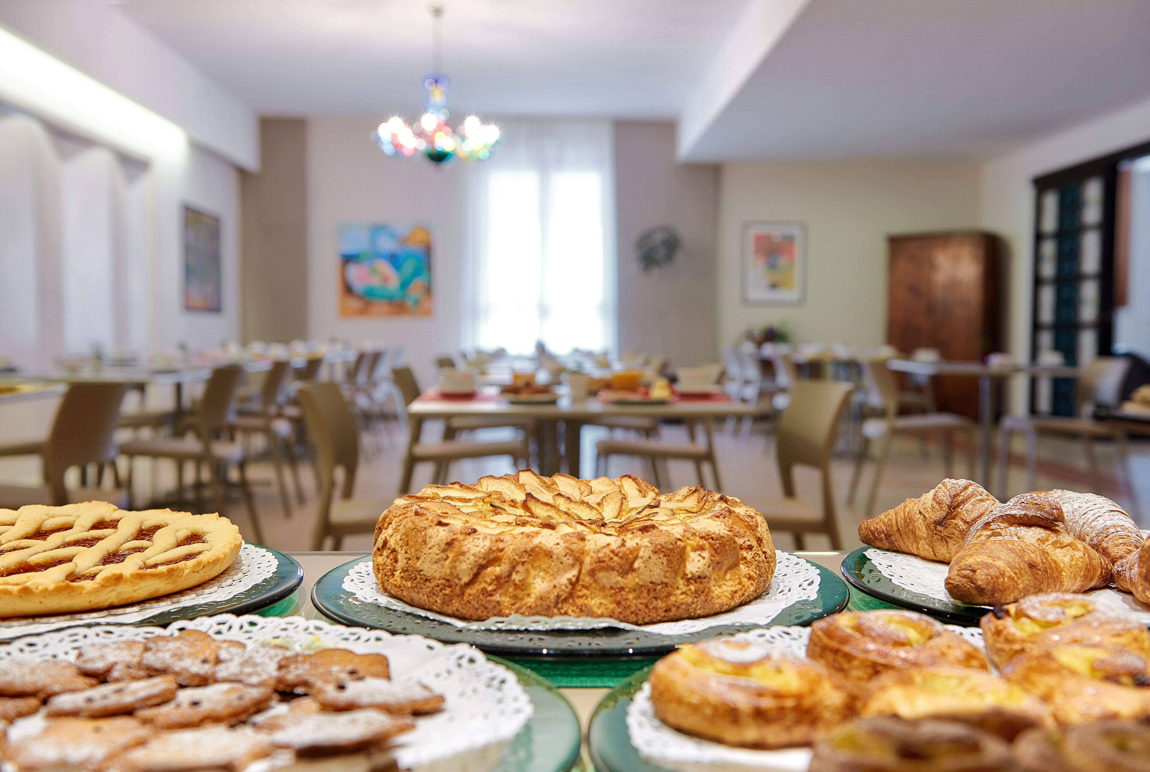 A dining area with a variety of pastries on display, including a lattice-topped pie, croissants, and other baked goods. Tables are arranged with chairs, and the room has a chandelier and artwork on the walls.