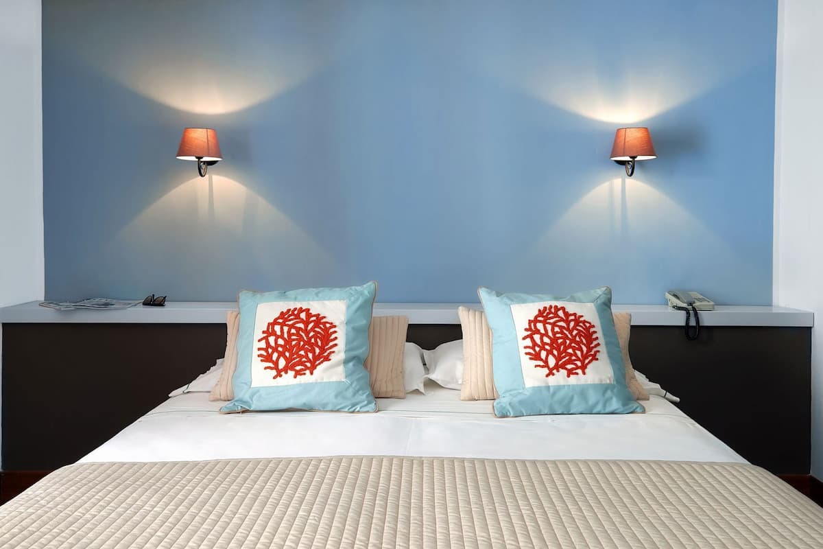 Une chambre d’hôtel comprenant un lit, deux lampes fixées au mur et deux coussins décoratifs à motifs coralliens.
