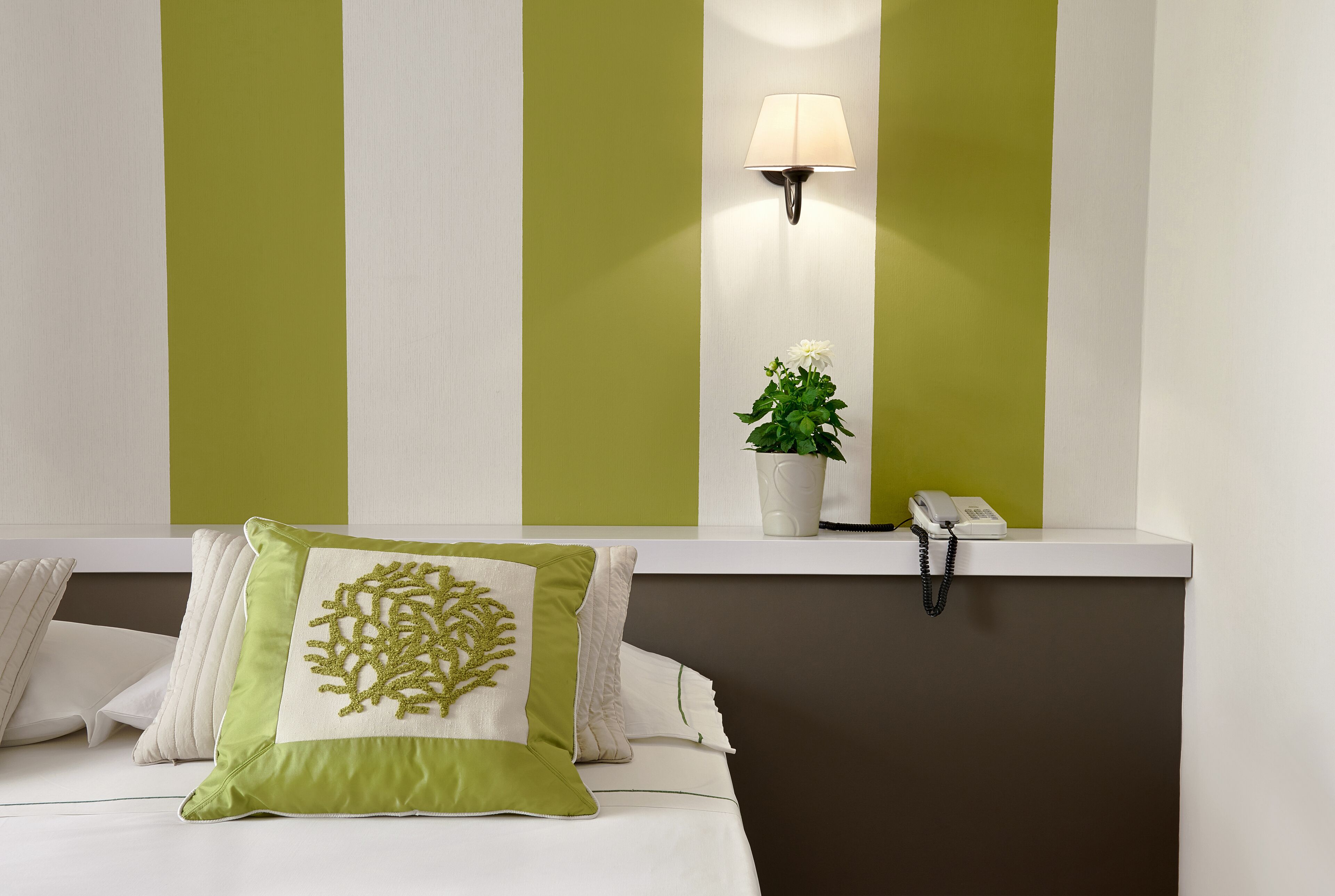 Habitación de hotel con cama, almohadas, mesita de noche con lámpara, teléfono y planta en maceta.