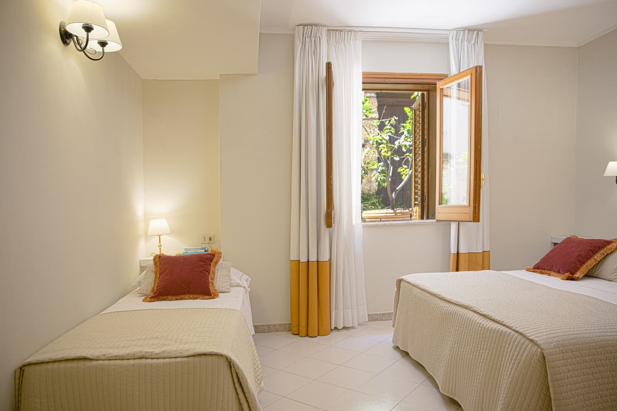 Une chambre d’hôtel avec deux lits, une fenêtre avec des rideaux, une lampe fixée au mur et une vue sur la verdure à l’extérieur.