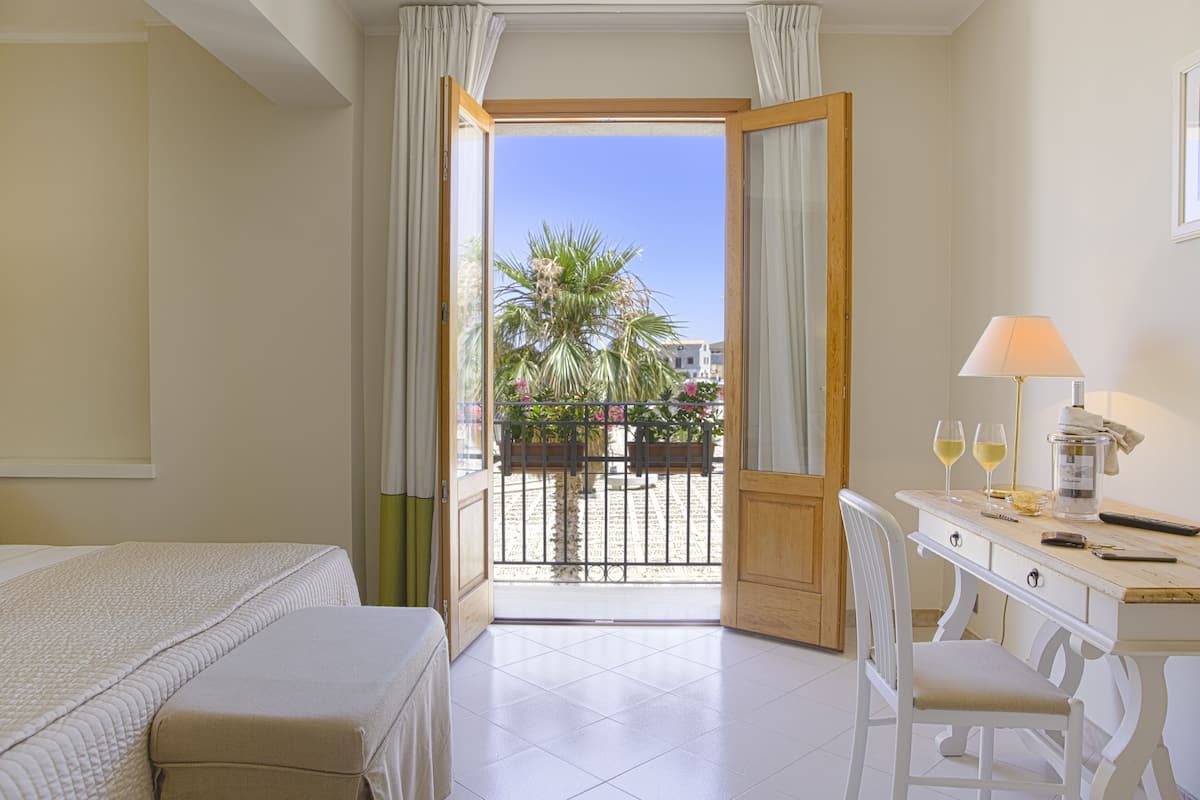 Une chambre d’hôtel avec un lit, un bureau doté d’une lampe, deux verres de vin et une vue sur un palmier et un balcon.