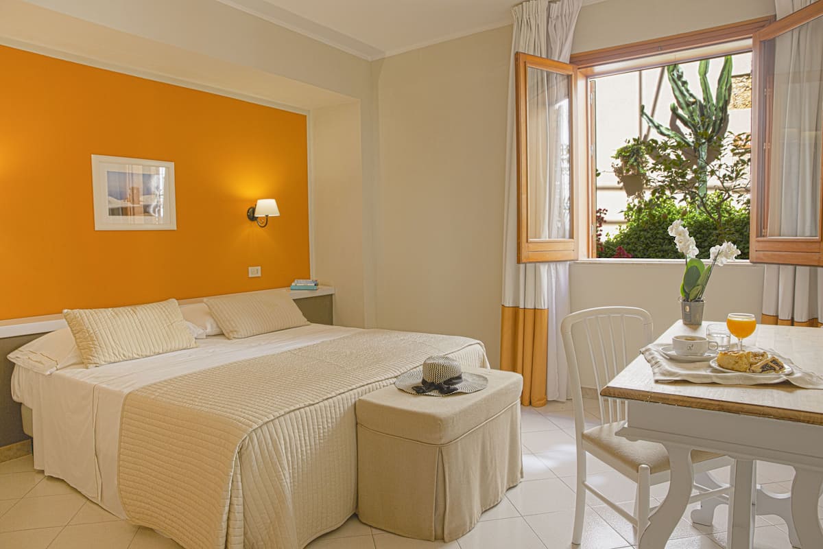 Une chambre d’hôtel avec un lit, une table à manger et une chaise, une fenêtre avec des rideaux et une plante en pot.