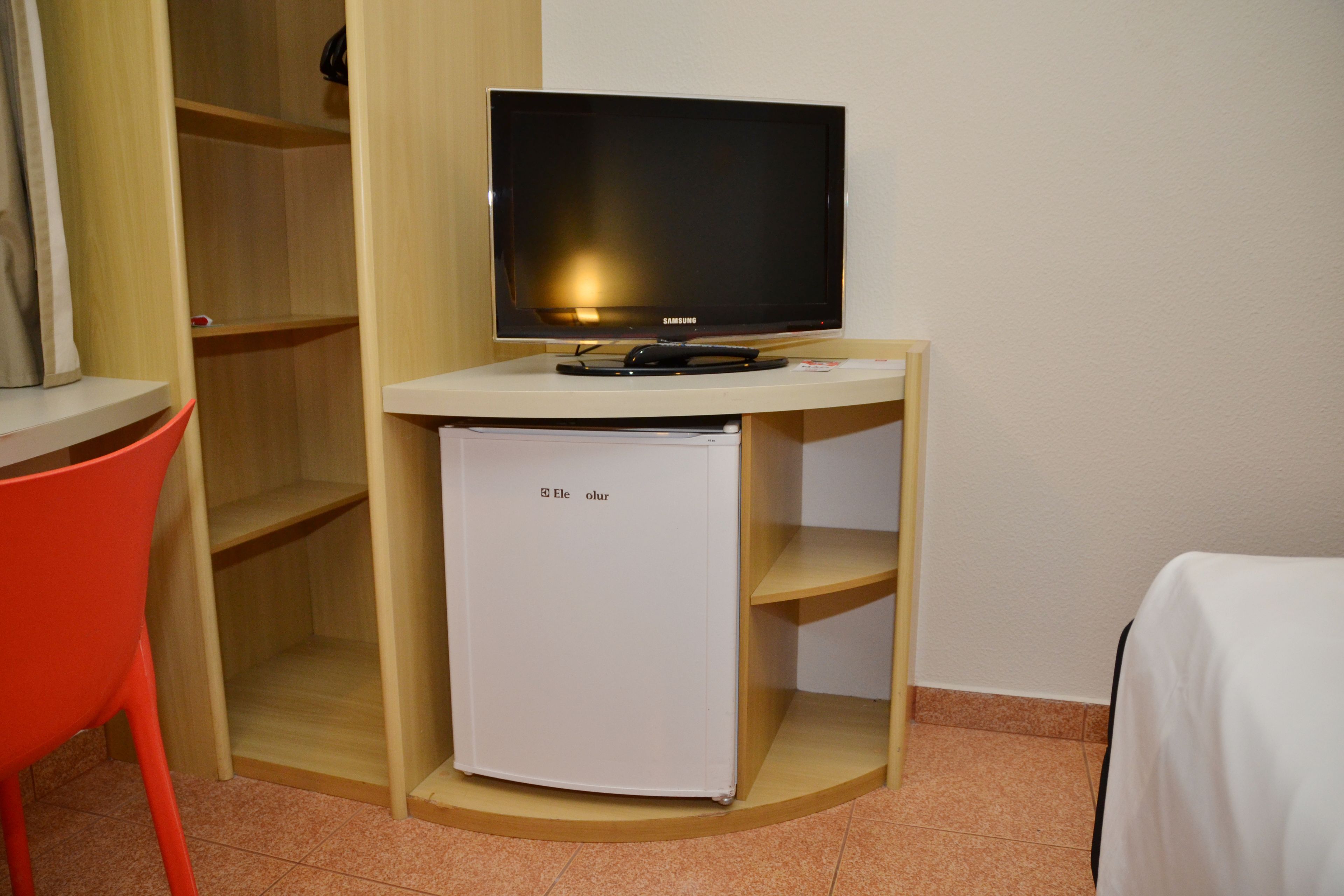 A compact room with a television on a wooden stand, a small white refrigerator, and a red chair.