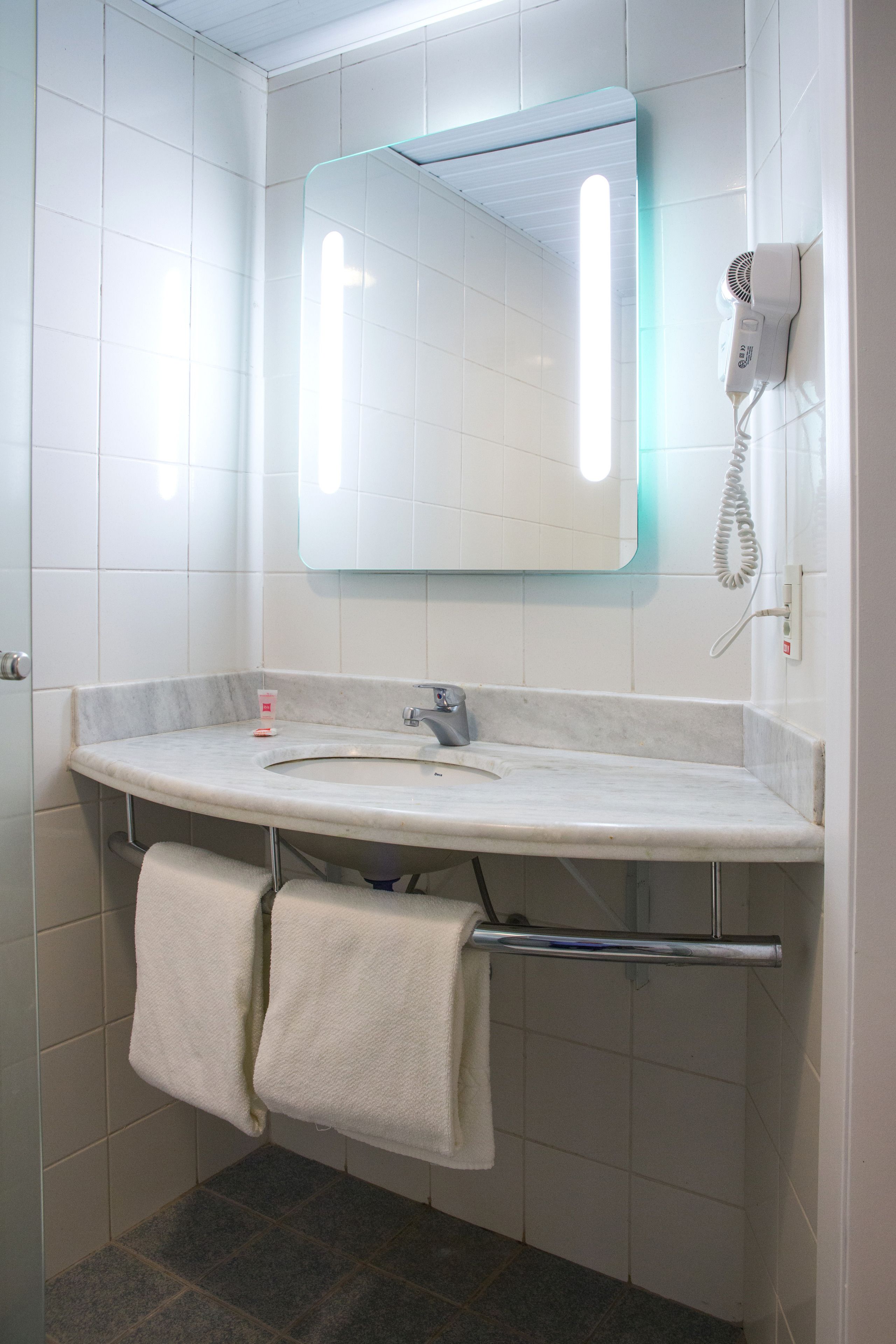 A bathroom with a white tiled wall, a large mirror, a white sink, and a hairdryer on the wall.