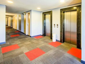 A hotel corridor with carpeted flooring, red carpet runners, and two elevators with marble surrounds. - ibis Aracatuba (Aracatuba)
