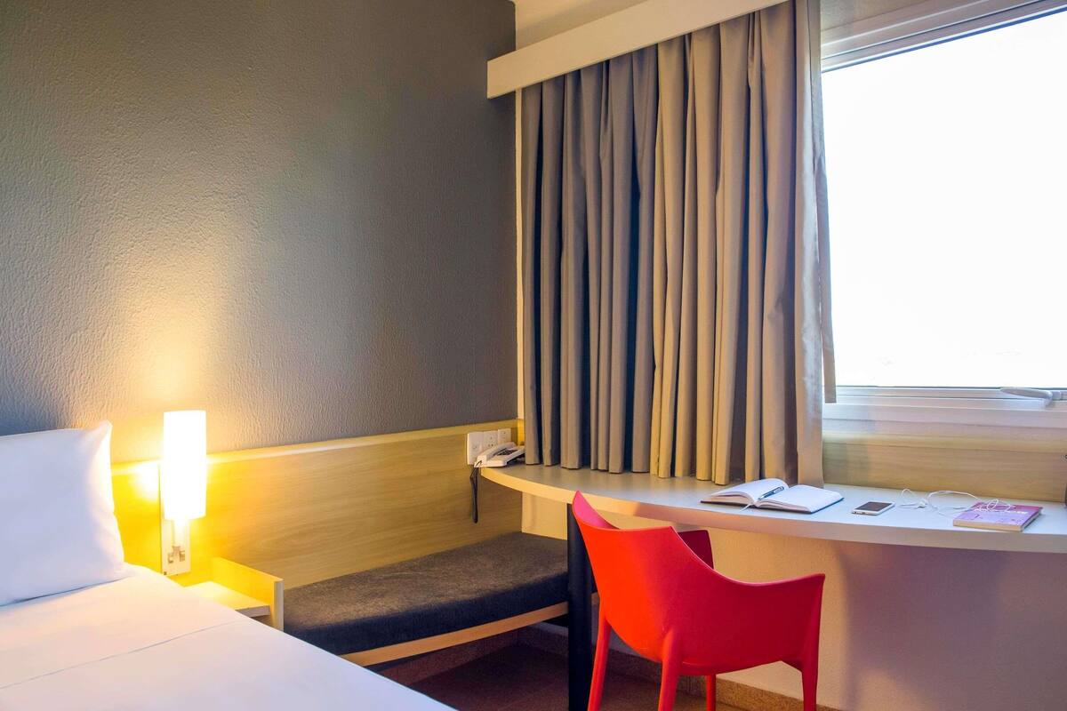 A hotel room with a bed, bedside lamp, desk, chair, and a window with curtains.