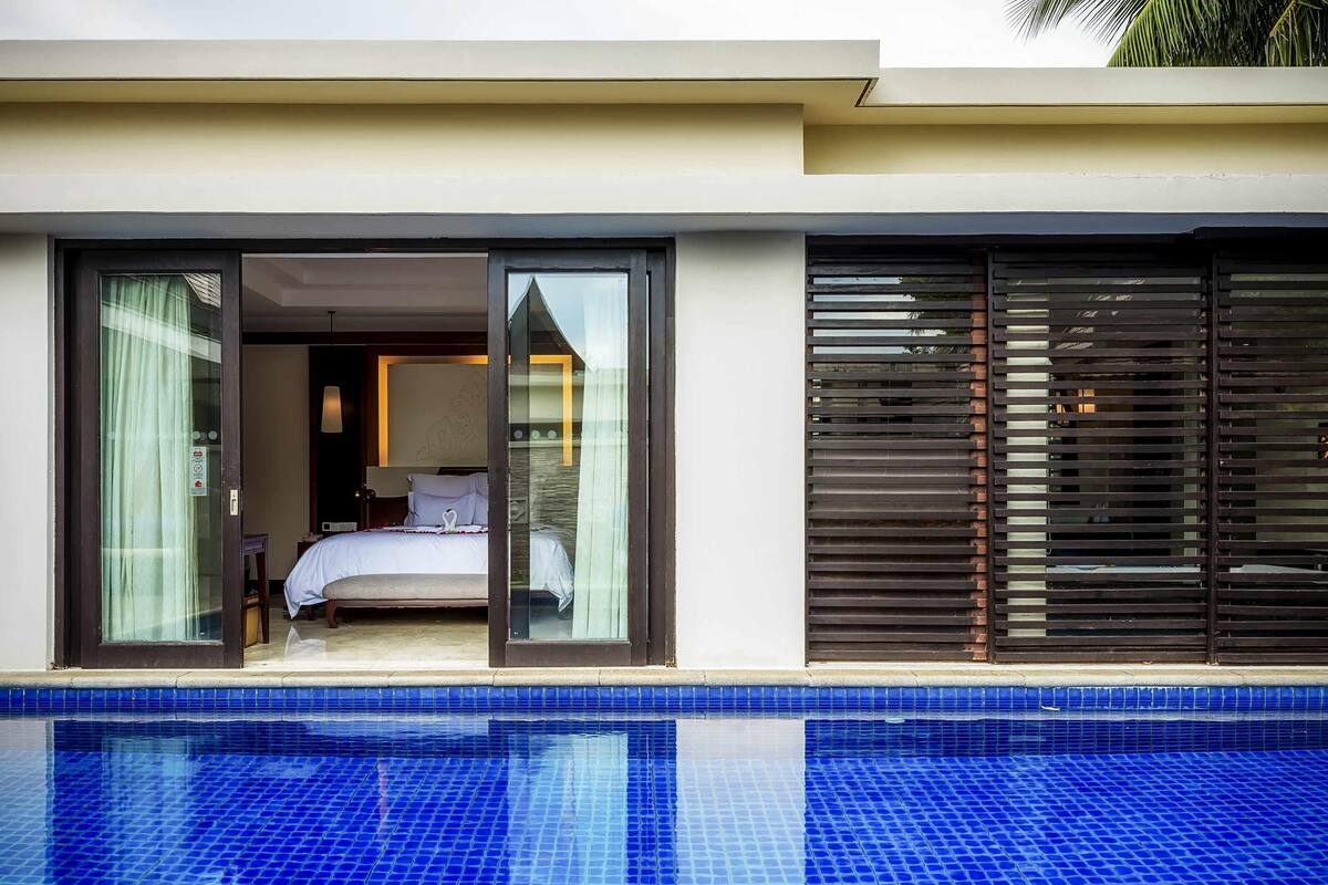 A modern hotel room with a glass door leading to a balcony, a pool with blue tiles, and a bed with white linens.