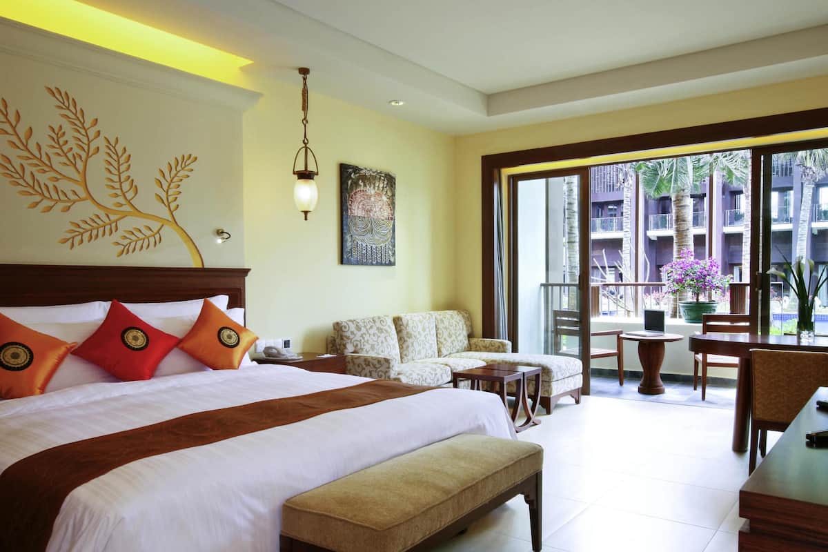 A hotel room with a large bed, a sofa, a dining table, and a balcony with a view of the surrounding buildings.