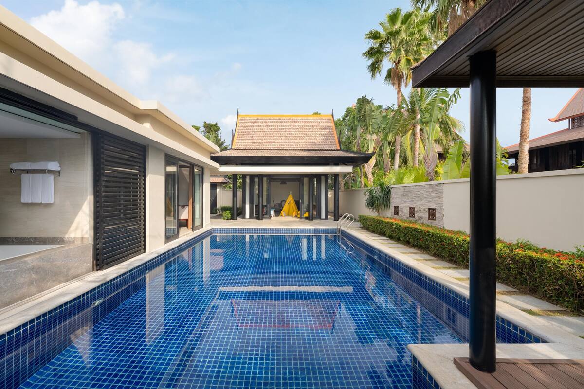 A swimming pool with a tiled deck, a covered area, and a view of a building with a tiled roof.