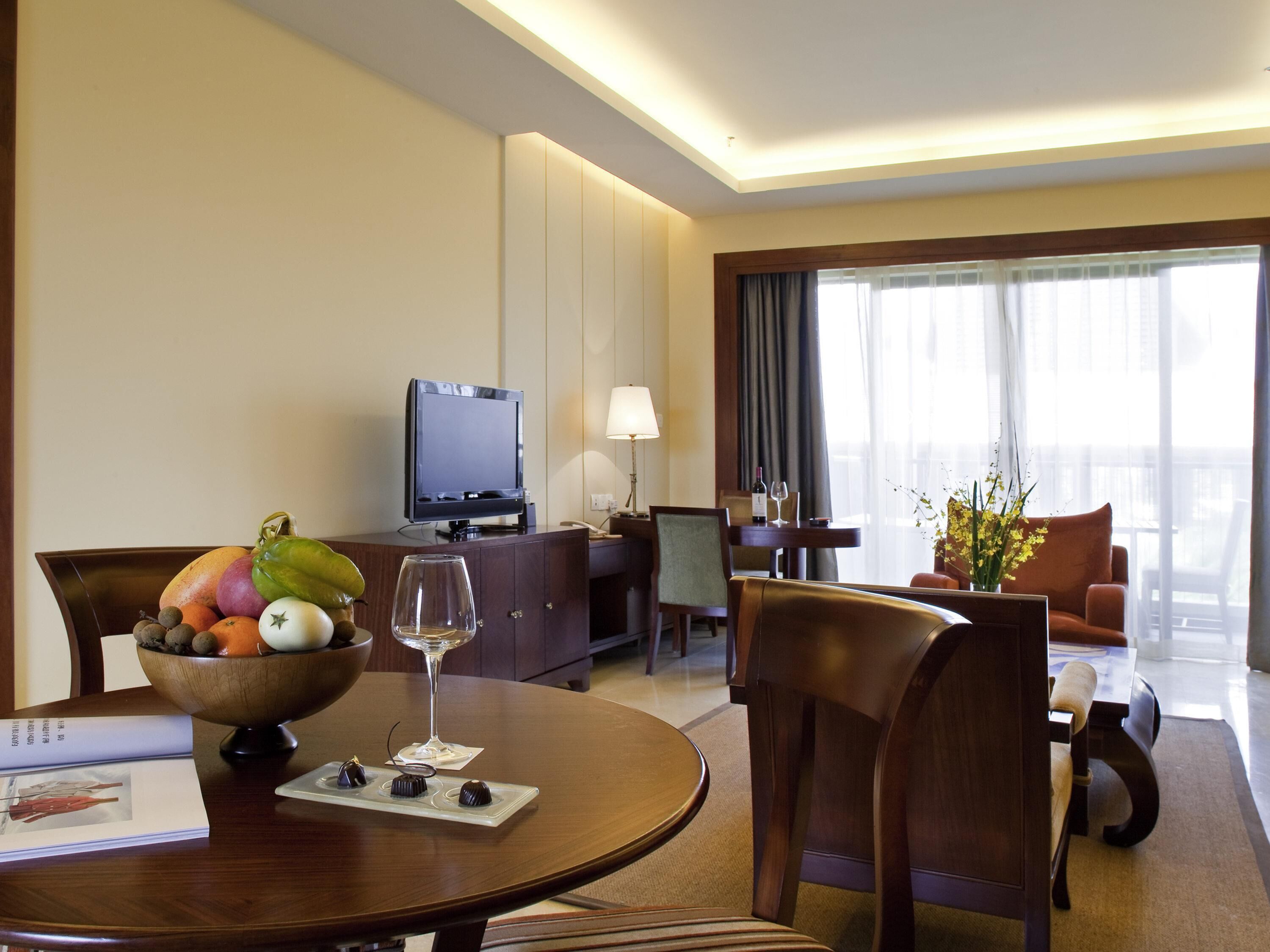 A hotel room with a dining table, chairs, a TV, a sofa, and a balcony with a view.