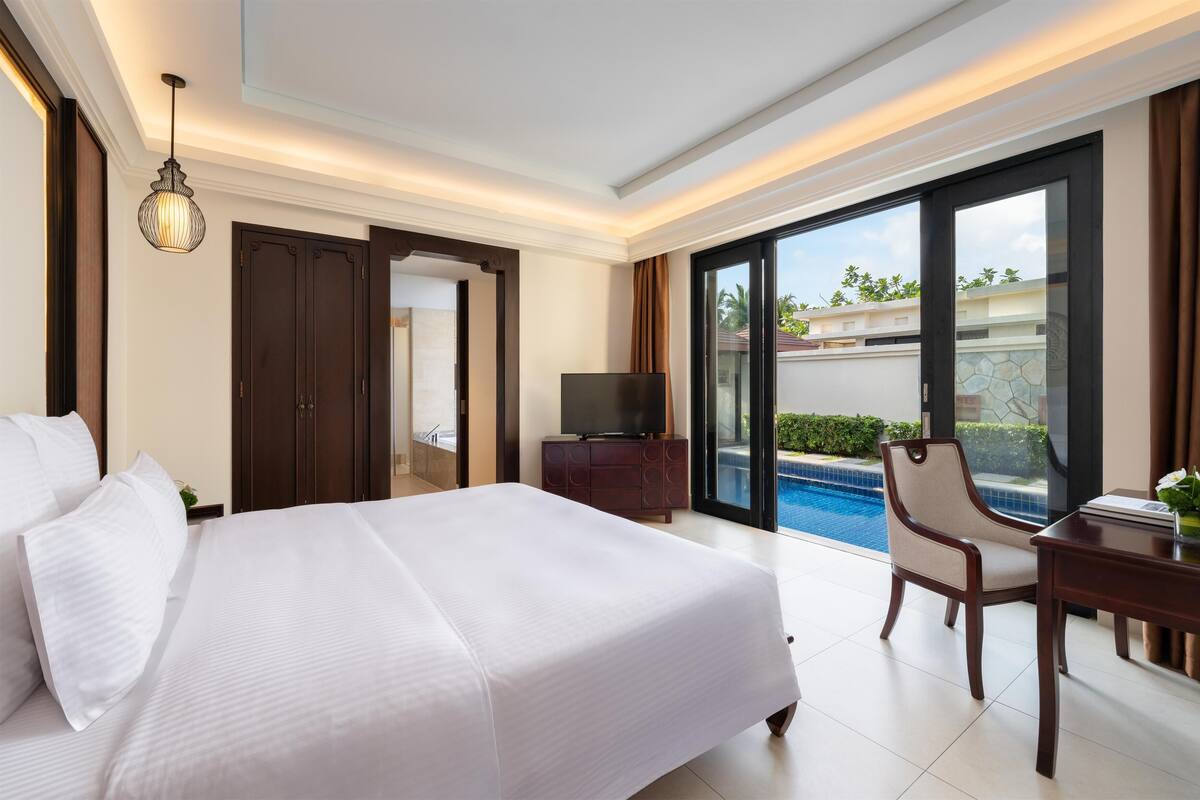 A hotel room with a bed, a TV, a desk, a chair, and a view of a swimming pool.