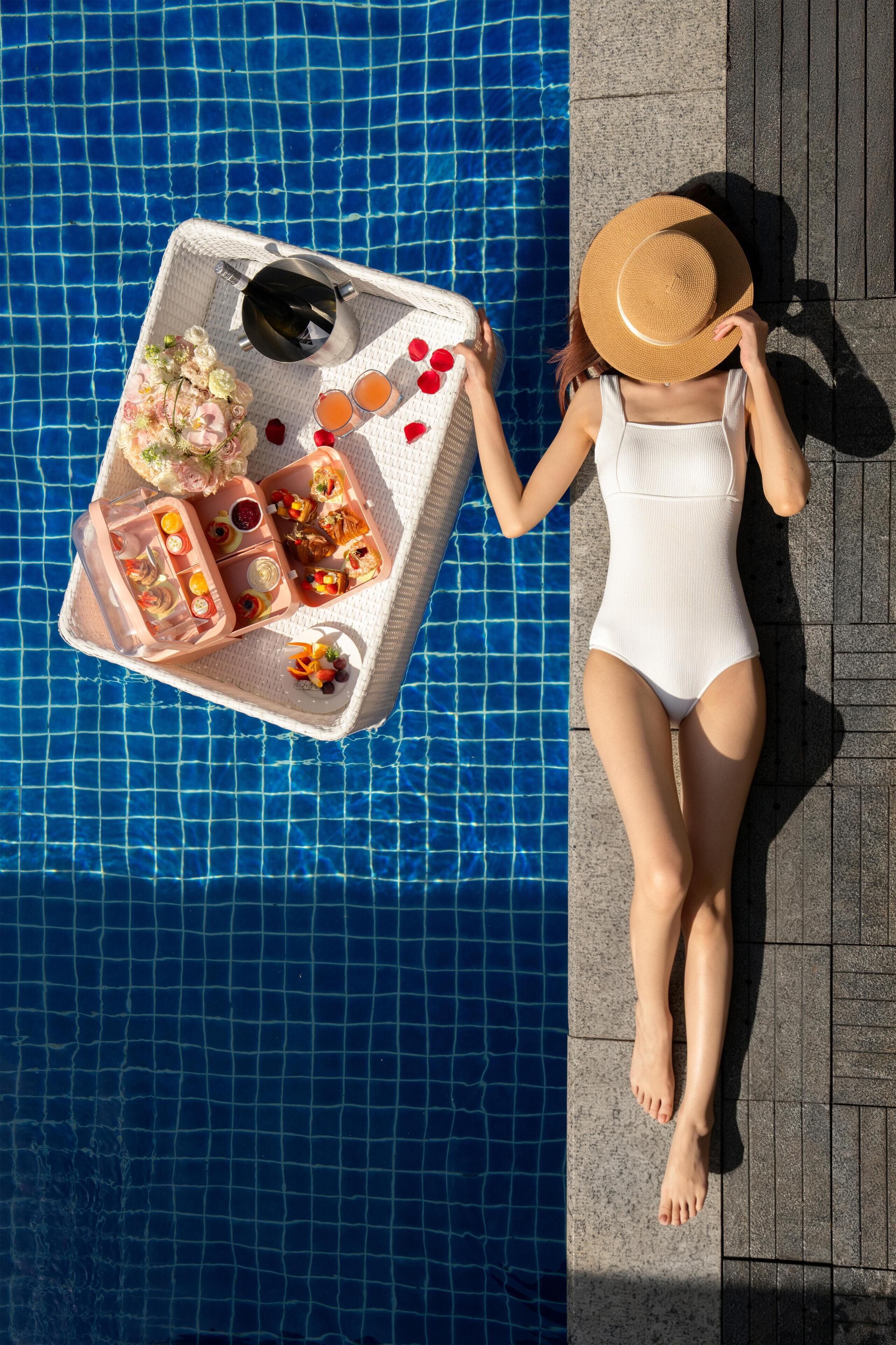 A person lying on a poolside lounge chair with a tray of food and drinks.