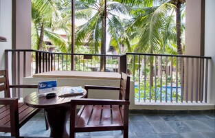 A balcony with a wooden table and chair, a sofa, and a view of palm trees and a swimming pool.
