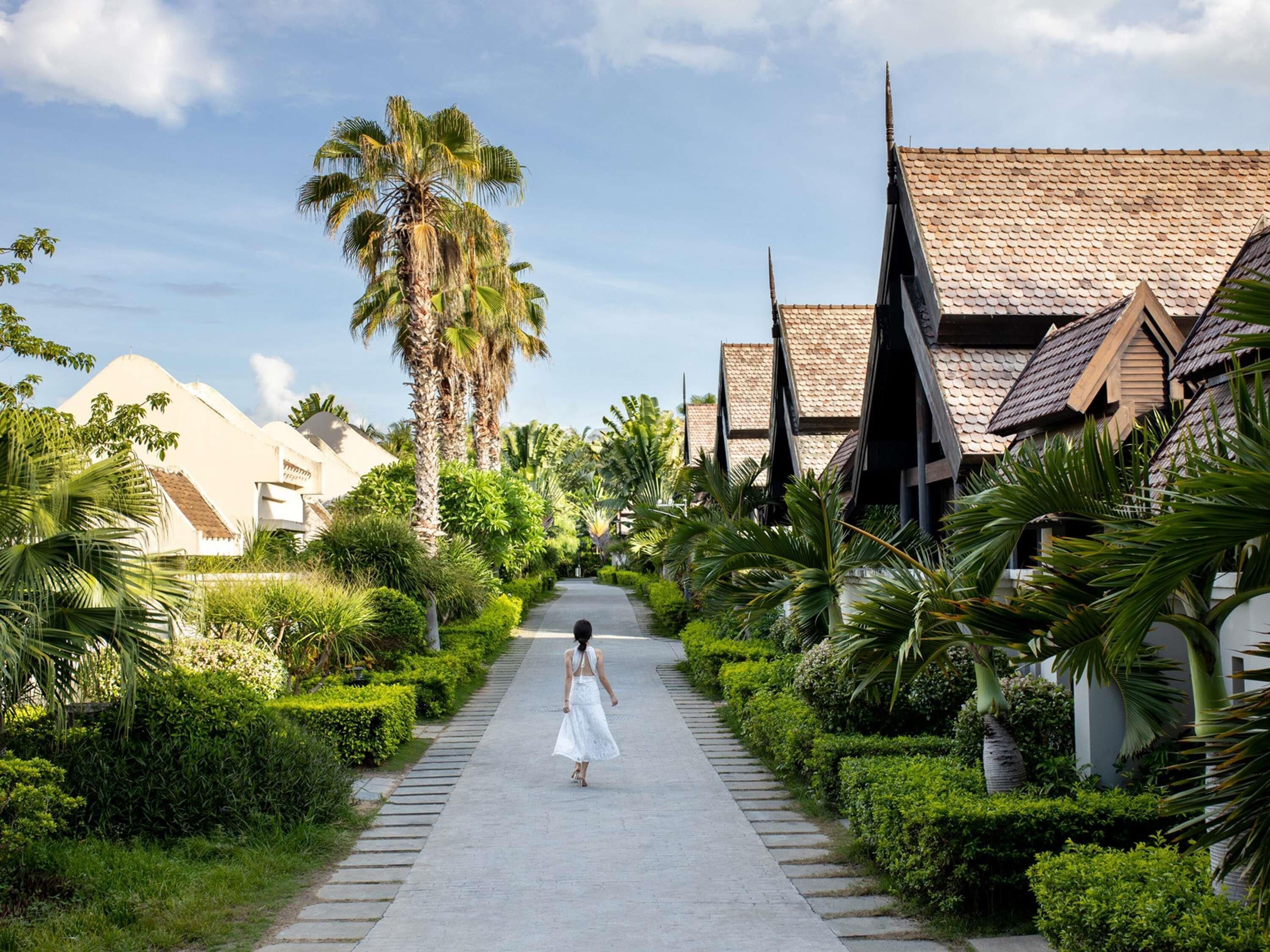 A pathway lined with lush greenery and traditional architecture, leading to a building with a tiled roof.