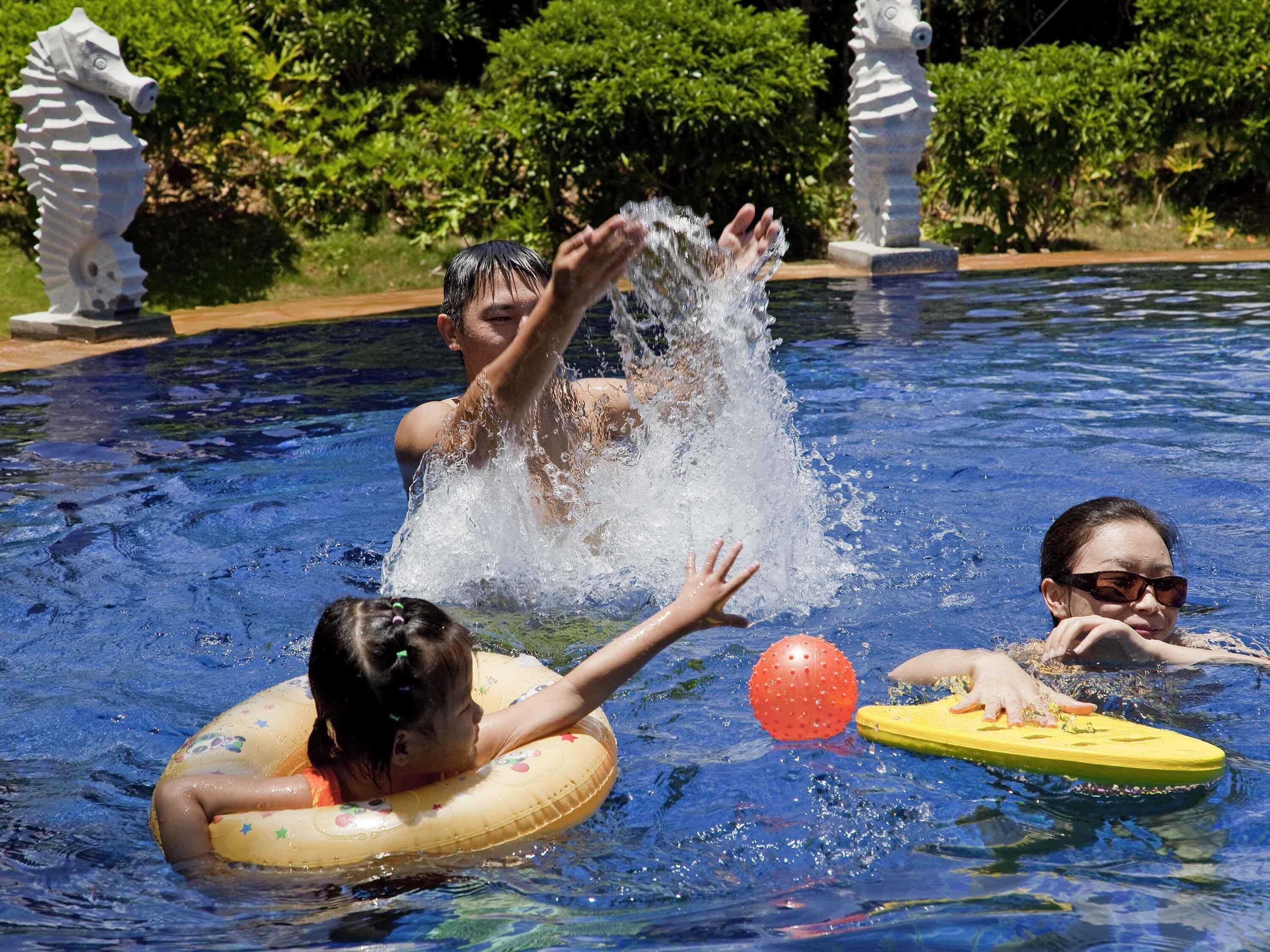 Piscina con dos personas y un niño en el agua, rodeada de follaje y estatuas decorativas.