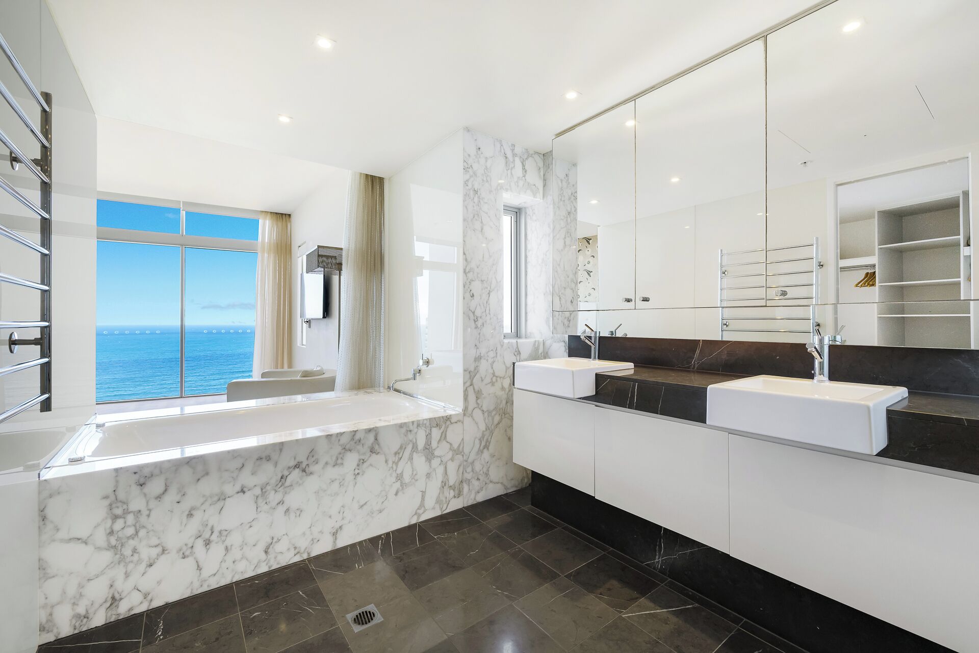 A modern bathroom with a large window offering a sea view, a bathtub, and two sinks with marble countertops.