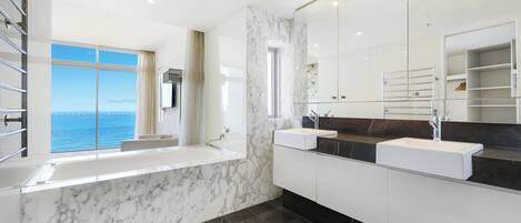 A modern bathroom with a large window offering a sea view, a bathtub, and two sinks with marble countertops.