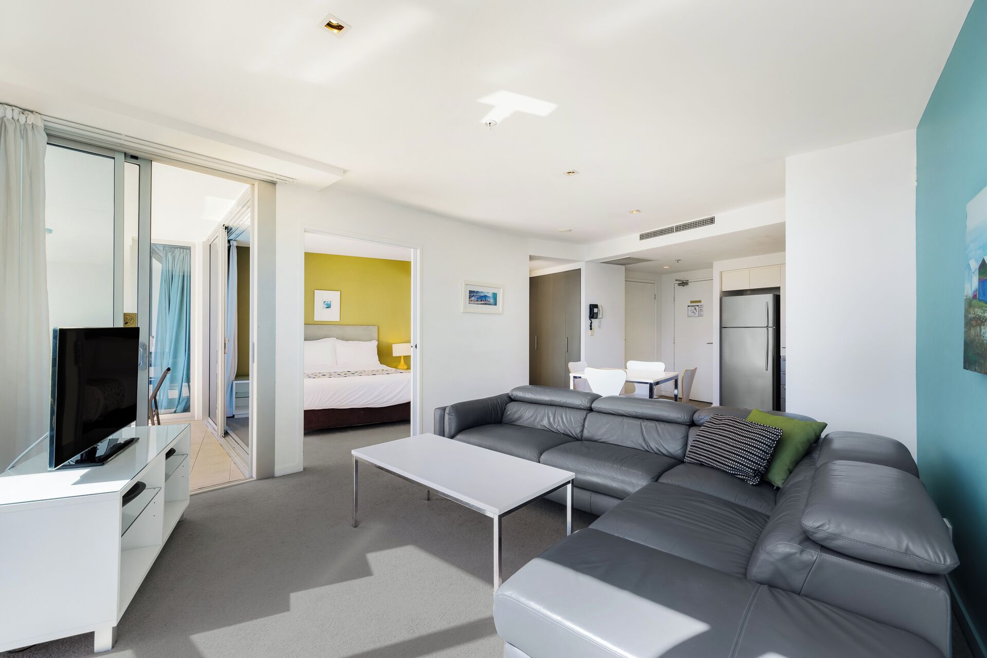 Una habitación de hotel moderna con un sofá gris, una mesita blanca pequeña, un televisor y una cama frente a una pared amarilla.