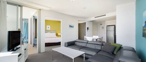 Una habitación de hotel moderna con un sofá gris, una mesita blanca pequeña, un televisor y una cama frente a una pared amarilla.
