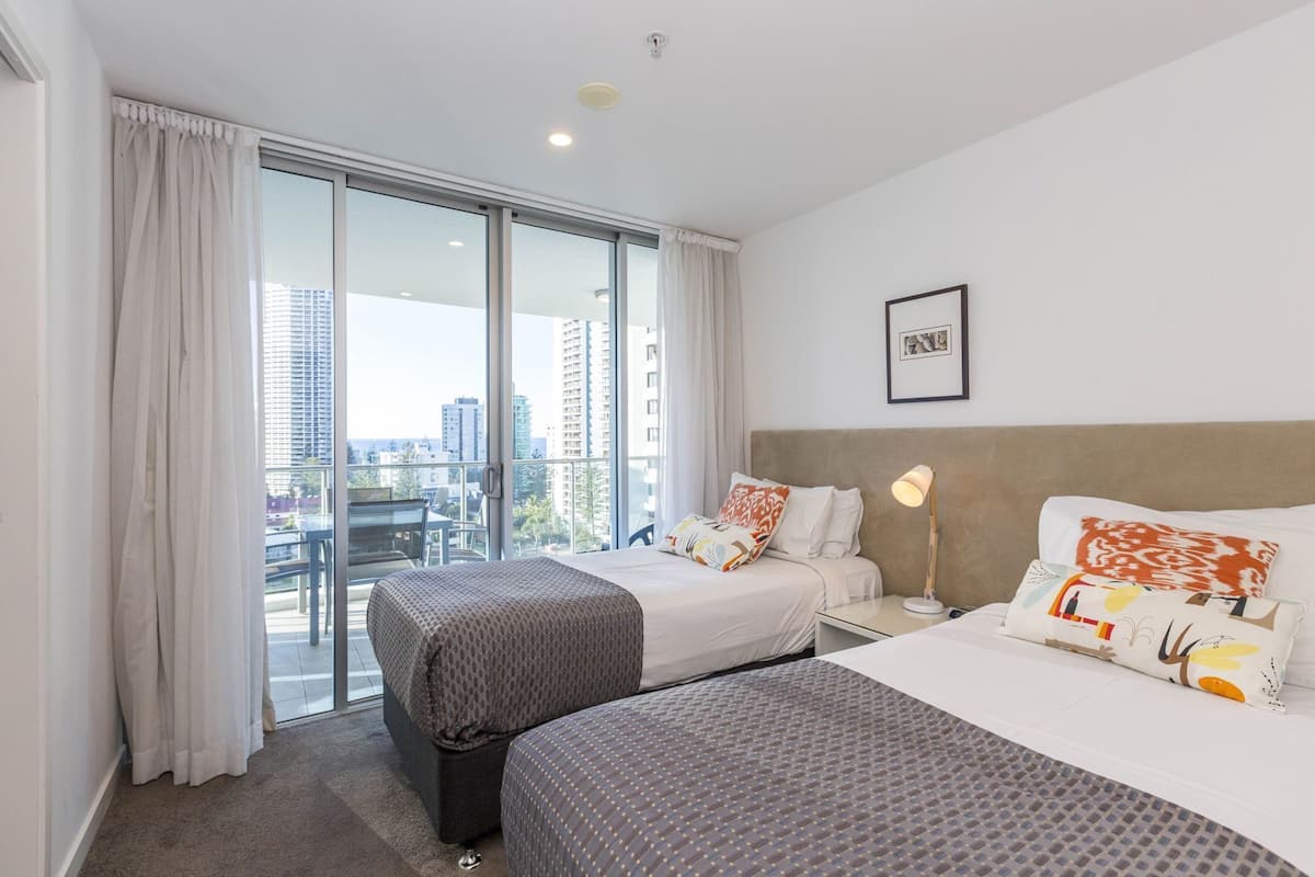 Een hotelkamer met twee bedden, een groot raam met gordijnen, uitzicht op de stad, een lijst aan de muur en een bedlampje.