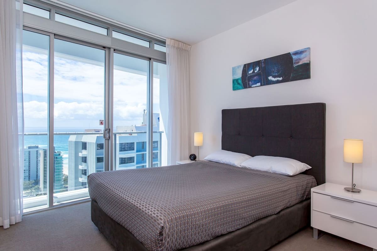 Een hotelkamer met een groot bed, een nachtkastje, een lamp, een schilderij en uitzicht op de stad en de oceaan.