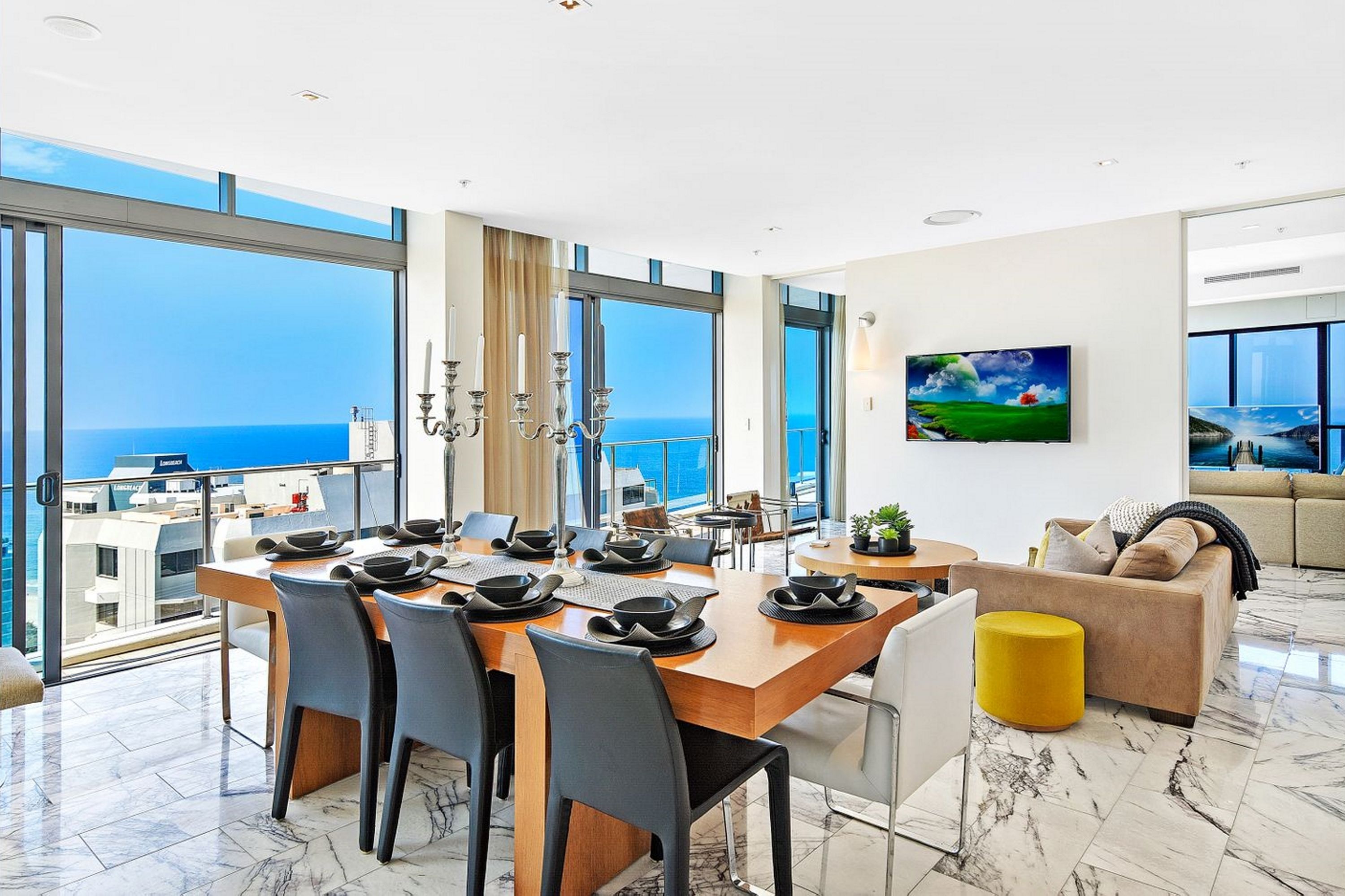 Un espace de repas moderne avec une grande table dressée pour six personnes, de grandes fenêtres offrant une vue sur la mer et un coin salon confortable avec un canapé et un pouf jaune.