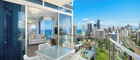 A balcony with a glass table and chairs, overlooking a cityscape with high-rise buildings and a clear sky.