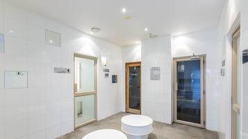 Un baño moderno con azulejos blancos, dos taburetes blancos redondos, una puerta de cristal e indicaciones de servicios de sauna y vapor.